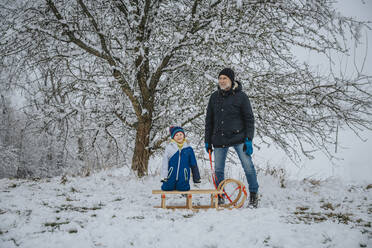 Father and son standing with sled in front of tree during winter - MFF07634