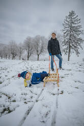 Lächelnder Vater betrachtet den Jungen, der neben dem Schlitten im Schnee liegt - MFF07639