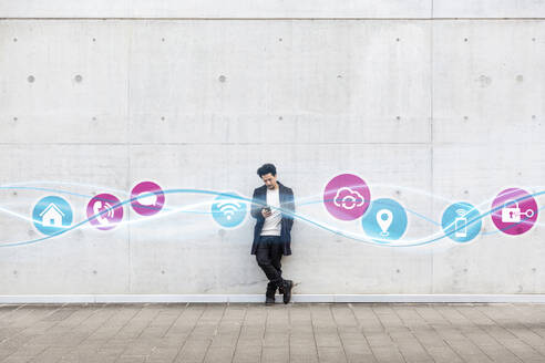 Young man using mobile phone while standing with icons on glowing wave pattern on footpath - EIF00624