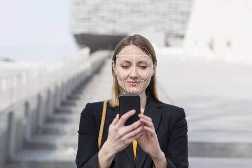 Businesswoman unlocking mobile phone with face recognition technology while standing outdoors - EIF00634