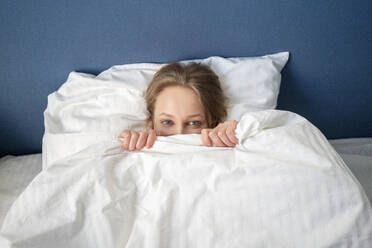 Young woman peeking while holding blanket at home - VPIF03740