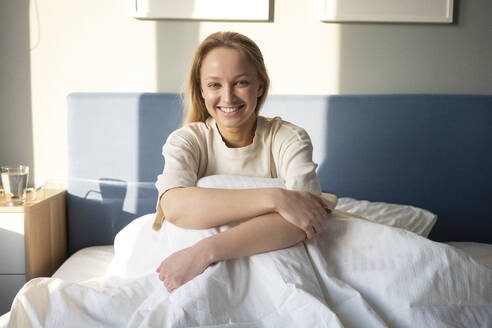 Junge Frau lächelt, während sie die Knie auf dem Bett zu Hause umarmt - VPIF03744