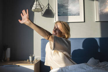 Smiling woman stretching hand toward sunlight while sitting on bed at home - VPIF03746