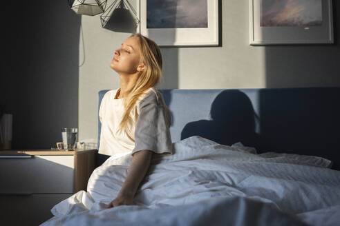 Sonnenlicht auf einer Frau, die mit geschlossenen Augen auf einem Bett zu Hause sitzt - VPIF03747