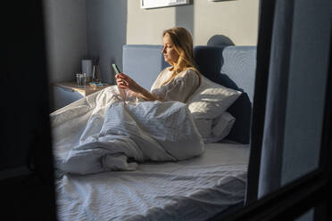 Woman using mobile phone while sitting with blanket on bed at home - VPIF03762