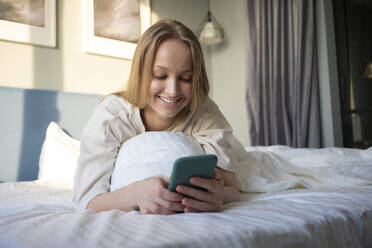Lächelnde Frau, die ein Mobiltelefon benutzt, während sie auf dem Bett liegt - VPIF03763