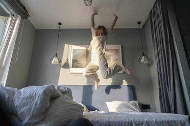 Carefree young woman with hand raised jumping on bed at home - VPIF03764