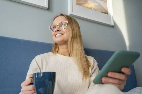 Blonde Frau schaut weg, während sie zu Hause mit Kaffeetasse und Mobiltelefon sitzt - VPIF03768