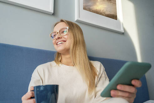 Junge Frau hält Kaffeetasse und Mobiltelefon und lächelt zu Hause - VPIF03769