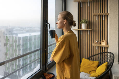 Frau schaut durch ein Fenster, während sie mit einer Kaffeetasse zu Hause steht - VPIF03782