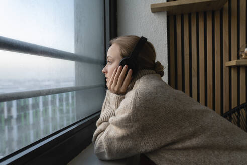 Frau hört Musik über Kopfhörer, während sie durch das Fenster zu Hause schaut - VPIF03786