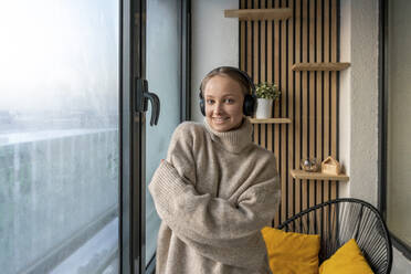 Junge lächelnde Frau mit Kopfhörern, die mit verschränkten Armen vor einem Fenster zu Hause steht - VPIF03788