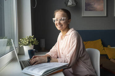 Smiling woman wearing eyeglasses sitting by laptop at home - VPIF03791