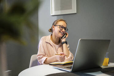 Smiling woman with laptop and book talking on mobile phone at home - VPIF03797