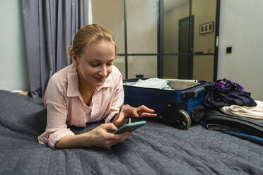 Junge lächelnde Frau, die ein Smartphone benutzt, während sie zu Hause auf dem Bett liegt - VPIF03811