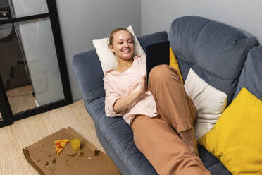 Woman using digital tablet while resting by food on sofa at home - VPIF03820