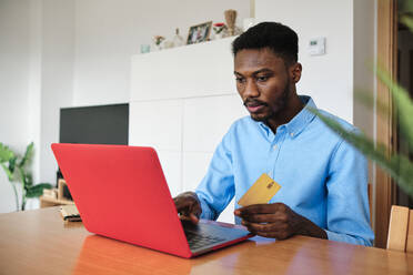 Geschäftsmann, der eine Kreditkarte hält, während er zu Hause einen Laptop benutzt - AGOF00087