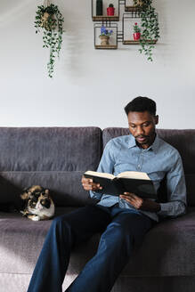 Handsome man reading book while sitting by cat in living room - AGOF00095