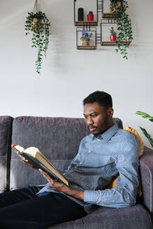 Man reading book while sitting on sofa at home - AGOF00096
