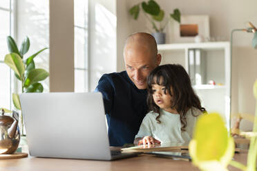 Vater arbeitet am Laptop, während er mit seinem Kleinkind im Home Office sitzt - SBOF03303
