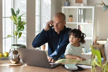 Father talking on mobile phone while sitting with daughter in home office - SBOF03305