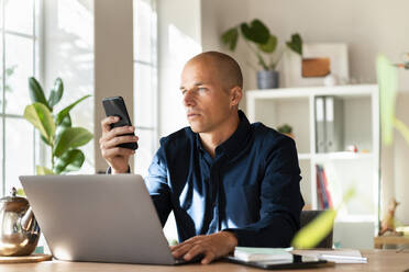Männlicher Unternehmer arbeitet am Smartphone, während er zu Hause am Laptop sitzt - SBOF03306