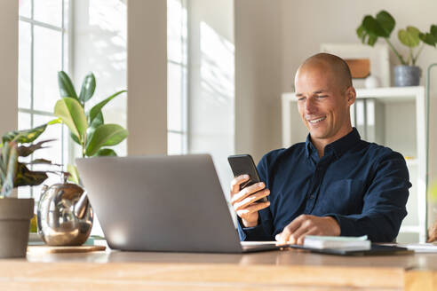 Lächelnder Geschäftsmann mit Laptop und Mobiltelefon bei der Arbeit zu Hause - SBOF03307