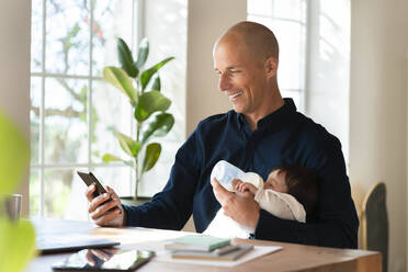 Vater füttert ein kleines Mädchen mit Milch, während er im Büro zu Hause am Smartphone arbeitet - SBOF03308