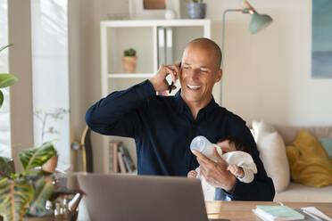 Lächelnder Vater, der mit seinem Smartphone telefoniert, während er seiner Tochter zu Hause Milch gibt - SBOF03309