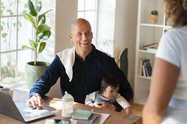 Lächelnder Mann sitzt mit Baby und sieht Frau bei der Arbeit zu Hause an - SBOF03310