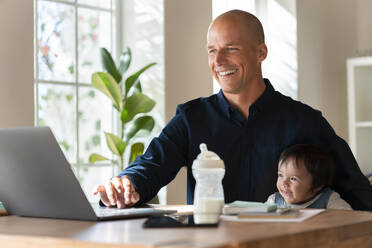 Happy father and daughter looking at laptop while working in home office - SBOF03311