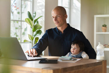 Männlicher Unternehmer arbeitet am Laptop, während er sein Baby im Home Office hält - SBOF03312