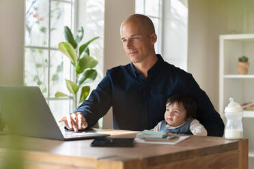 Male entrepreneur working on laptop while holding baby in home office - SBOF03312