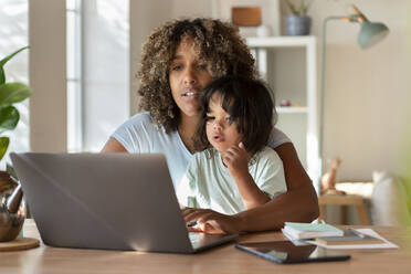 Mother and toddler daughter looking at laptop together while working from home - SBOF03315