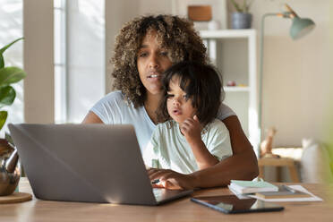 Mutter und kleine Tochter schauen gemeinsam auf den Laptop, während sie von zu Hause aus arbeiten - SBOF03315