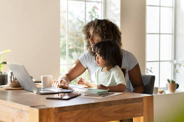 Mother working on laptop while toddler looking at it in home office - SBOF03316