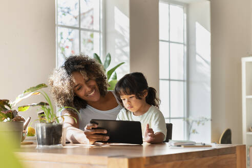 Lächelnde Mutter und Tochter schauen auf ein digitales Tablet, während sie im Wohnzimmer sitzen - SBOF03319