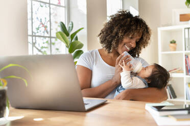 Smiling mother feeding milk to baby while sitting with laptop in home office - SBOF03321