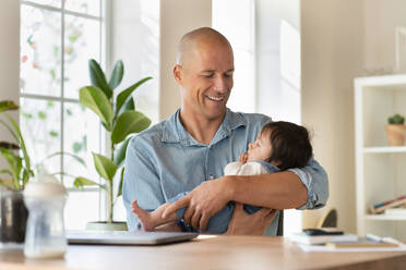 Lächelnder Vater, der sein Baby im Wohnzimmer hält und anschaut - SBOF03328