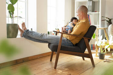 Vater sieht sein schlafendes Baby an, während er zu Hause auf einem Sessel sitzt - SBOF03338
