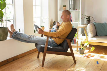 Smiling father using digital tablet and holding baby while sitting on armchair at home - SBOF03339