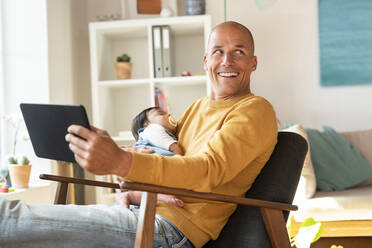 Smiling father looking away while holding sleeping daughter and digital tablet in living room - SBOF03340