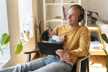 Lächelnder Vater hört Musik, während er ein schlafendes Baby und ein digitales Tablet im Wohnzimmer hält - SBOF03353