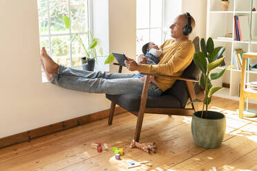 Mid adult father looking away while listening music and holding sleeping baby at home - SBOF03355