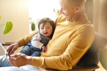 Vater hält digitales Tablet und lächelndes Baby, während er zu Hause auf einem Sessel sitzt - SBOF03360