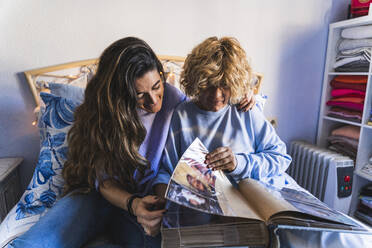 Lächelnde Tochter schaut sich ein Fotoalbum an, während sie mit ihrer Mutter im Schlafzimmer sitzt - MPPF01596