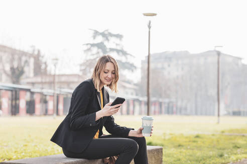 Mid adult businesswoman with coffee disposable cup using mobile phone at park - EIF00661