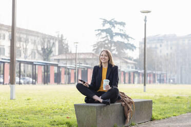 Smiling businesswoman sitting with mobile phone and disposable cup at park - EIF00662