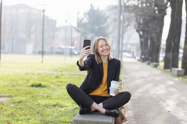Cheerful businesswoman taking selfie through mobile phone while sitting on bench at park - EIF00665