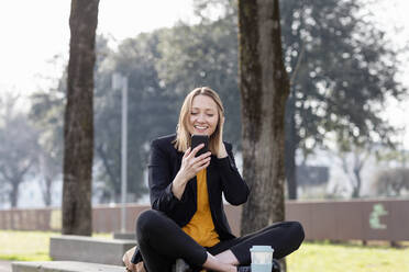 Lächelnde Geschäftsfrau, die ein Smartphone benutzt, während sie im Schneidersitz im Park sitzt - EIF00667